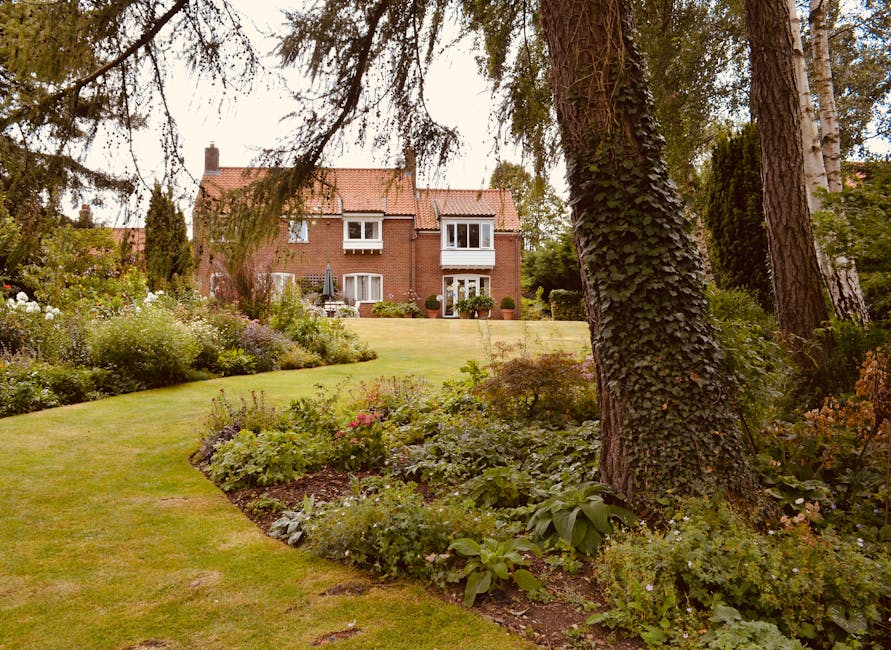 A well-maintained garden scene featuring neatly trimmed hedges and a variety of plants. In the foreground, there are low, dense hedges with fresh green foliage, alongside flowering plants with purple